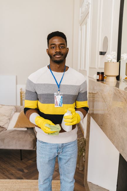 A professional cleaner from Croydon Carpet Cleaners stands in a well-lit living room, holding a spray bottle and a cleaning cloth. The room features a beige marble countertop with decorative vases and